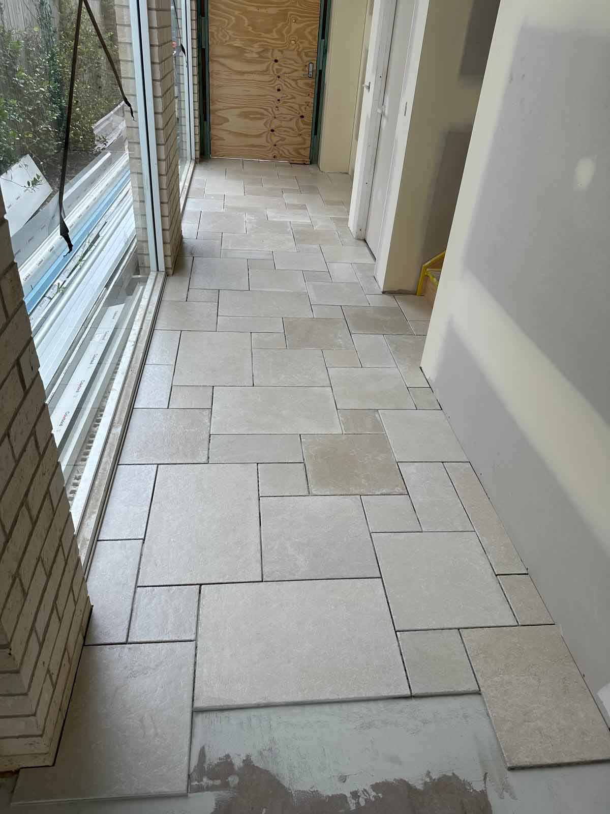 Hallway floor laid with cream tiles in a mixed-size pattern beside glass doors