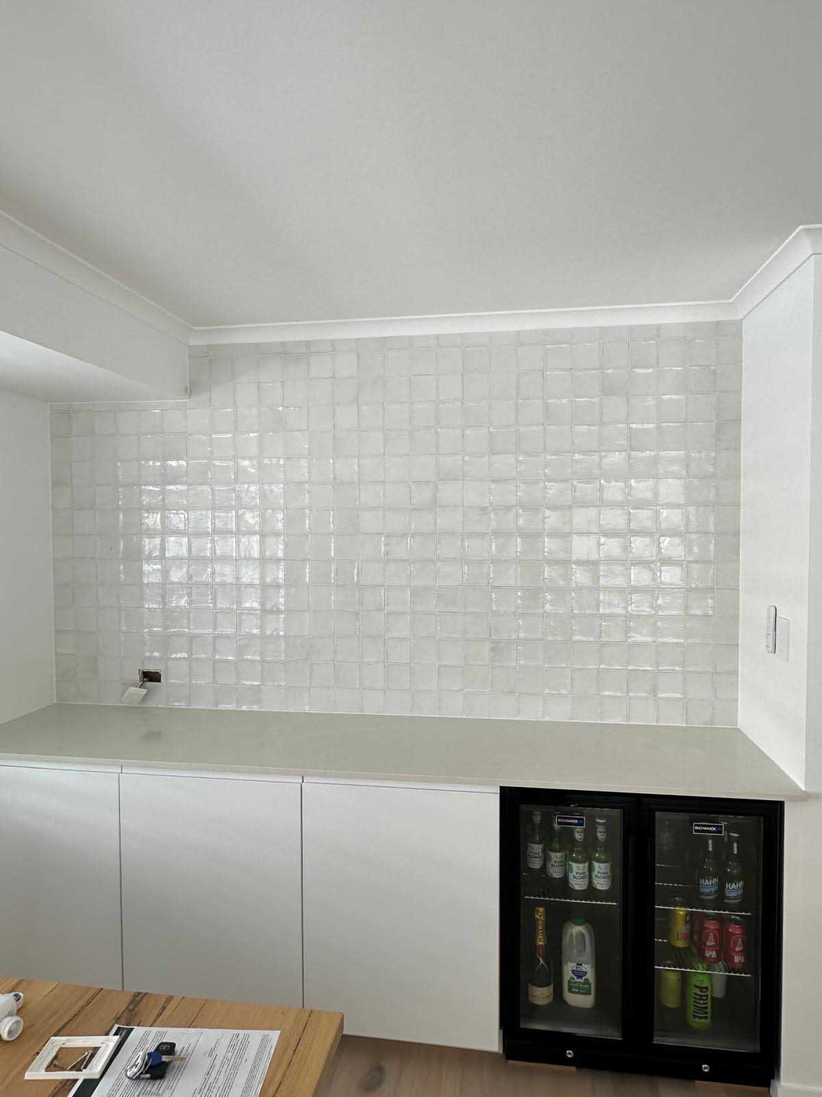 Glossy white square splashback tiles above white cabinets and a bar fridge